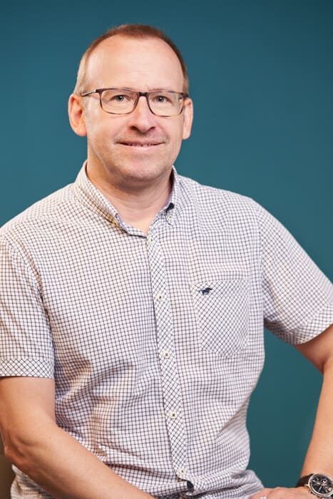 Porträt eines männlichen Mitarbeiters von Königs Umzüge vor blauem Hintergrund, freundlich lächelnd mit Brille und kariertem Hemd